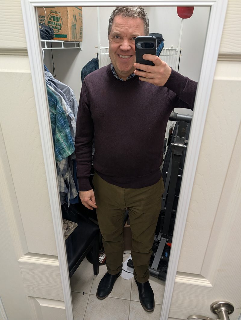 Mirror selfie of outfit being critiqued: burgundy v-neck sweater over plaid shirt, olive trousers, black Chelsea boots