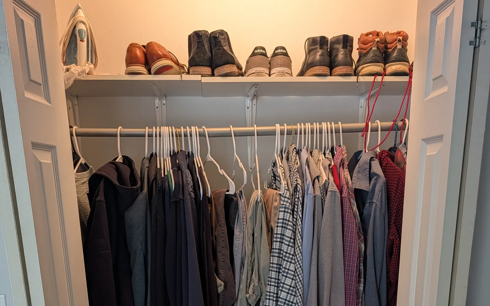 An open closet with shirts, sweaters and jackets on white hangers, and leather shoes, boots and sneakers lined up on the shelf above.