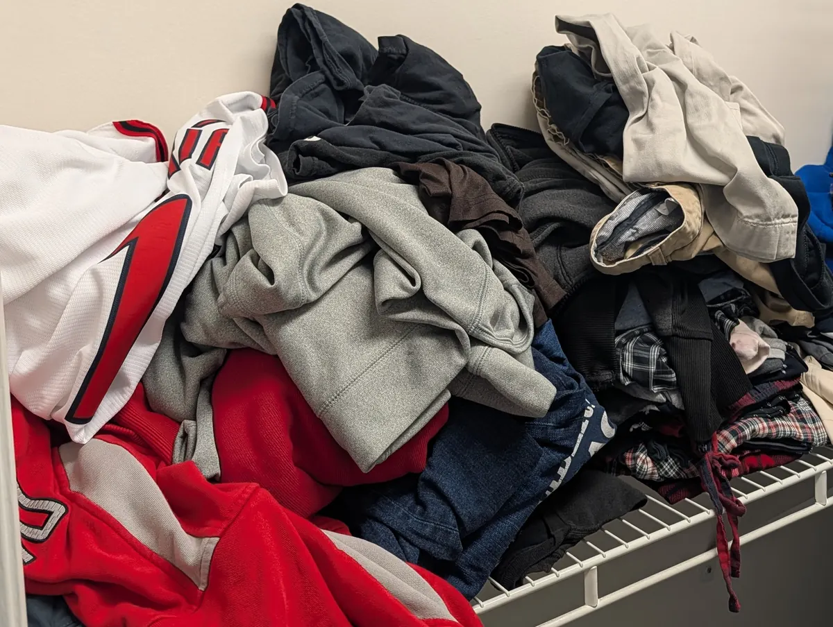 A heap of hoodies, jeans, flannels and a sports jersey piled on a wire closet shelf.
